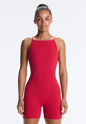 Žena stojaca čelom vpredu, oblečená v bezrukávový, jasne červený priliehavý overal s tenkými ramienkami a štvorcovým výstrihom.