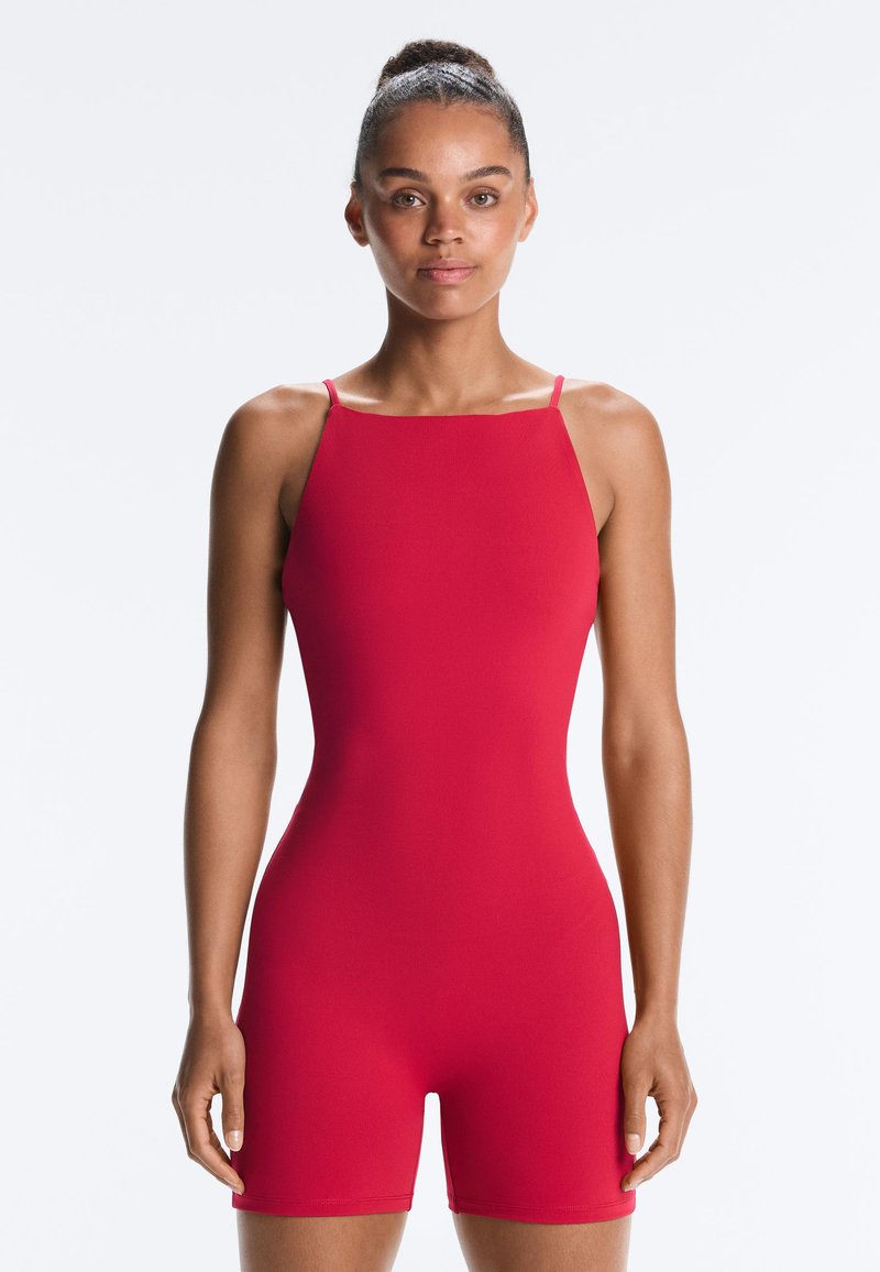 Femme debout, face à l'avant, portant une combinaison courte rouge vif sans manches, cintrée, avec des bretelles fines et un décolleté carré.
