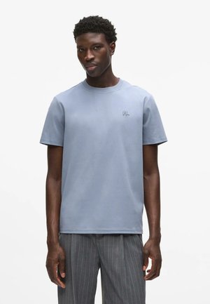 Jeune homme portant un t-shirt bleu clair à manches courtes et un pantalon gris à fines rayures, debout sur un fond blanc uni.