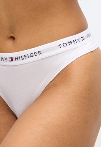 Balts kokvilnas tangas biksītes ar melnu un sarkanu joslu, uz kuras ir uzraksts "TOMMY HILFIGER". Gluda tekstūra, zems viduklis, piegulošs siluets.