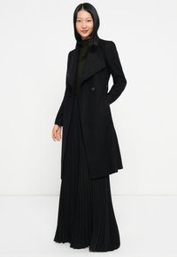 Casaco trench preto de lã com lapela larga, design de botão duplo e bolsos laterais, combinado com uma saia preta plissada.
