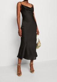 Zwarte satijnen midi-jurk met vallende halslijn, dunne bandjes, uitlopende zoom en een gladde textuur. Gecombineerd met versierde hakken en een kleurrijke tas.