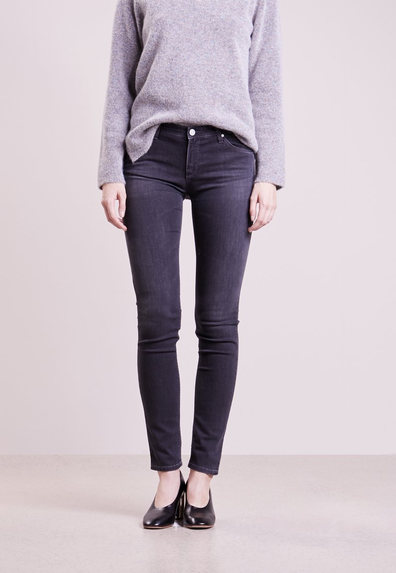 Czarne skinny jeans z metalowym guzikiem i zapięciem na zamek; połączone z szarym swetrem z dzianiny i czarnymi butami na wysokim obcasie.