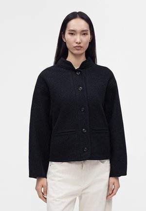 Femme aux longs cheveux foncés portant une veste noire à boutons et un pantalon blanc, debout devant un fond clair uni.
