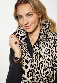Vest puf leopard cu imprimeu în nuanțe de crem și negru, având guler înalt, fermoar frontal și textură lucioasă; asortat cu un pulover negru.