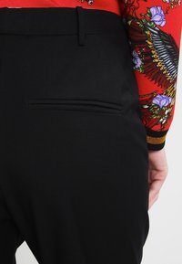 Svarta skräddarsydda byxor med en slät yta, med en enda bakficka och en slim fit. En blommig ärm är delvis synlig.