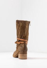 Brown suede boot with a mid-calf design, featuring a stacked wooden heel and a tan leather strap around the ankle. Zipper closure at the back.