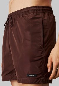Bañador burdeos con cinturilla elástica y cordón de ajuste. Presenta un bolsillo lateral y un logo negro de Calvin Klein en el dobladillo.