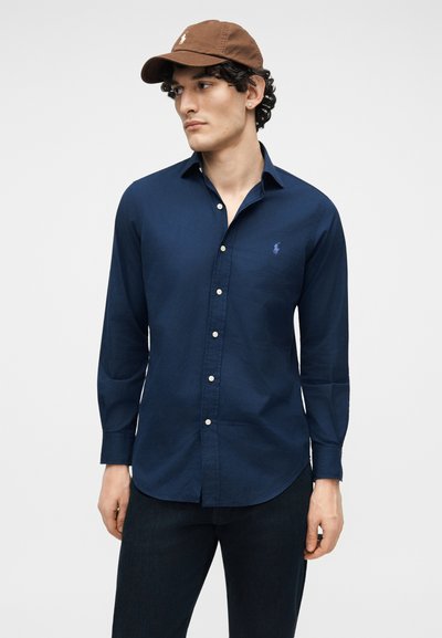 Muž oblečený v tmavomodrej košeli na gombíky a tmavých nohaviciach, pozerá sa bokom, so hnedou baseballovou čiapkou a náušnicami.