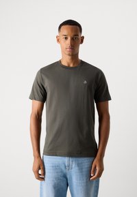Khaki green cotton T-shirt with crew neck, short sleeves, and small white logo on the chest. Worn with light blue denim jeans.
