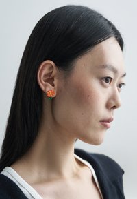 Oransje øredobber formet som blomster med grønn stilkdetalj, laget av et glatt materiale. Plassert mot en nøytral bakgrunn.