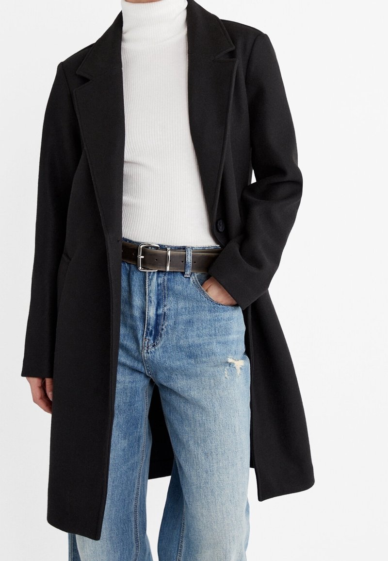 Persoon in een zwarte lange jas over een witte coltrui, gestopt in hoge taille blauwe jeans met een zwarte riem, met één hand in de zak.