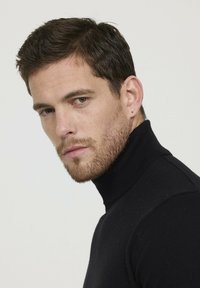 Pull noir à col roulé en matériau doux, design ajusté, col côtelé, porté par un homme regardant légèrement sur le côté devant un fond uni.