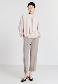 Blusa rosa chiaro con davanti drappeggiato e maniche a sbuffo, abbinata a pantaloni beige mirati e tacchi slingback neri a punta.
