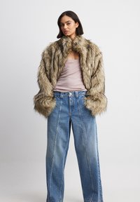 Lažni krzneni plašč v svetlo rjavi barvi z temnimi črtami, s visokim ovratnikom in širokimi rokavi, v kombinaciji s svetlo modrimi širokimi kavbojkami.