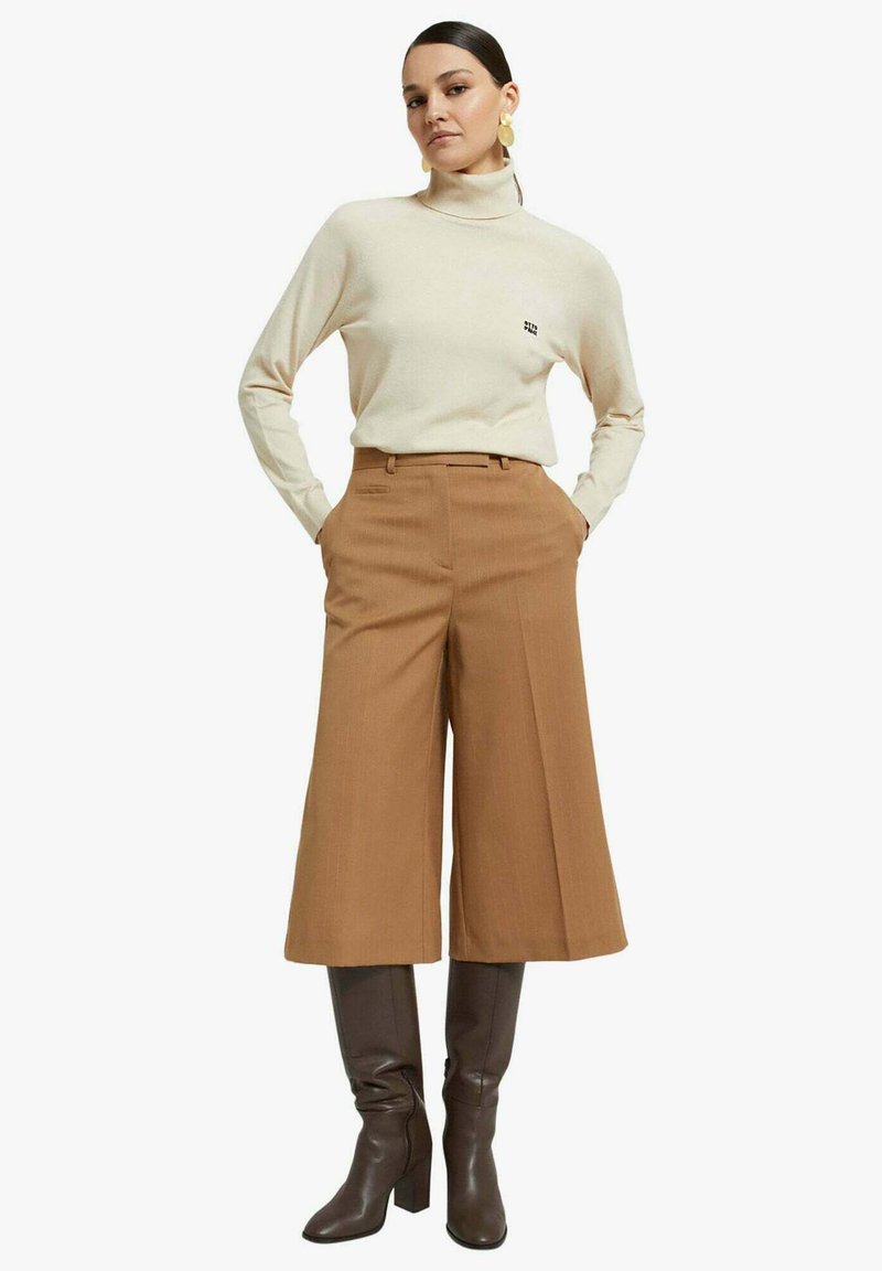 Maglione a collo alto beige, pantaloni marroni a gamba larga e stivali marroni al ginocchio. I pantaloni presentano dettagli con pieghe. Orecchini grandi a cerchio.