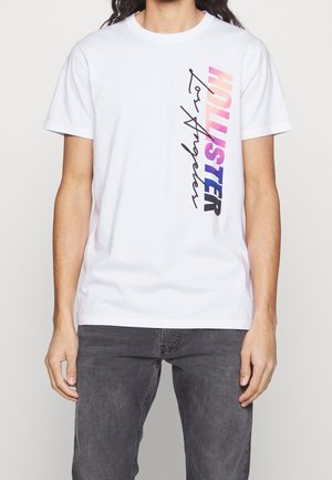 Mężczyzna w białym, krótki rękaw T-shircie z pionowym napisem "HOLLISTER" w gradiencie od różowego do niebieskiego oraz ozdobnym podpisem na lewej piersi, w połączeniu z szarymi dżinsami.