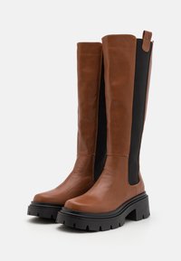 Paire de bottes hautes en cuir marron avec panneaux latéraux élastiques noirs et semelles épaisses en caoutchouc noir sur fond blanc.