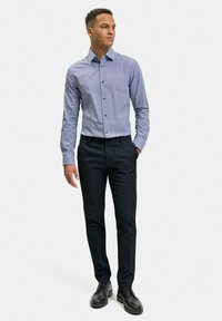 Blå mönstrad skjorta med knäppbar krage och långa ärmar, i kombination med mörka byxor och svarta skor, stående på en enfärgad bakgrund.