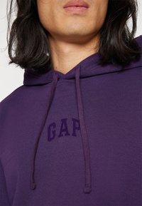 Sudadera morada hecha de una mezcla de algodón, con un bolsillo frontal, capucha con cordón y el logo "GAP" en morado más oscuro en el pecho.
