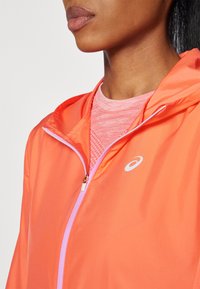Veste légère orange vif avec une fermeture éclair rose, arborant un logo sur la poitrine et une chemise texturée rose en dessous.