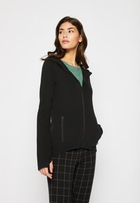 Hoodie noir zippé avec une capuche à cordon, poches latérales zippées et une texture lisse. Porté sur une chemise verte, associé à un pantalon à carreaux.