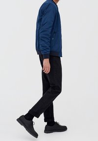 Námořnická modrá bomber bunda s texturovaným materiálem, černé slim-fit kalhoty a černé šněrovací boty, předváděno v bočním pohledu.