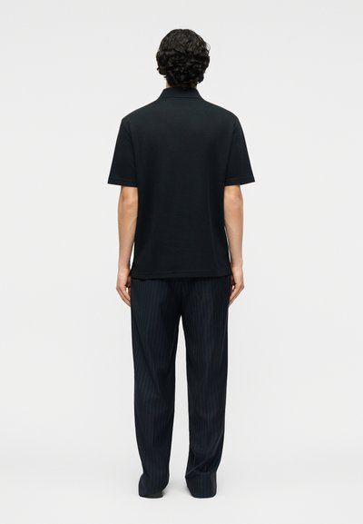 Άνδρας με σγουρά μαλλιά στέκεται με την πλάτη, φορώντας μαύρο κοντομάνικο πουκάμισο και σκούρο παντελόνι με λεπτές ρίγες, μπροστά σε απλό φόντο.