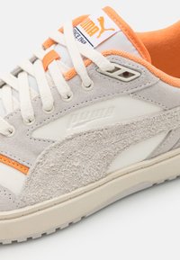 Gros plan sur une sneaker beige et orange Puma avec des détails en suède texturé, des lacets blancs et le logo Puma sur la languette et le côté.