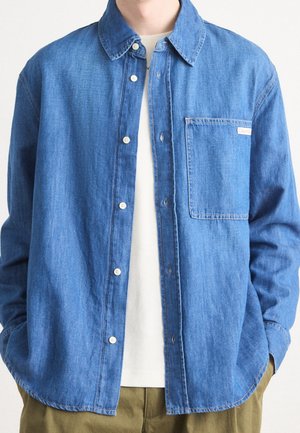 Person iført en åben blå denimskjorte over en hvid t-shirt med hænderne i khakifarvede bukselommer, beskåret over nakken.