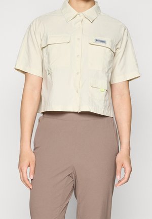 Osoba nosząca krótki beżowy T-shirt z krótkim rękawem z przednimi kieszeniami i brązowe spodnie z wysokim stanem.