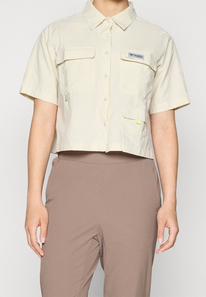 Personne portant une chemise courte beige à manches courtes avec des poches sur le devant et un pantalon marron taille haute.
