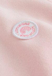 Svetloružová tkanina s hladkým povrchom obsahuje okrúhlu výšivku s obrázkom dúhy a textom v ružovej a bielej farbe.