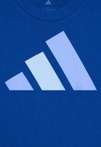 Modrá bavlnená tričko s veľkým bielym logom Adidas, ktoré obsahuje tri šikmé tvary. Hladká textúra a klasický dizajn s okrúhlym výstrihom.