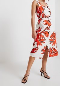 Robe midi florale avec une base blanche, ornée de motifs de feuilles rouges et noires vifs, avec un décolleté carré, une ceinture et une fente à l'avant.