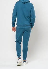 Tyrkysová mikina s kapucí a odpovídající joggers vyrobené z měkké tkaniny, s uvolněným střihem a žebrovanými manžetami. Tenisky s bílou základnou a námořnickými akcenty.