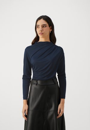 Top lungo navy a maniche lunghe con design drappeggiato sul davanti, abbinato a una gonna nera in pelle con dettagli cuciti e lunghezza maxi.