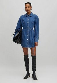 Denim jurk met lange mouwen, knoopsluiting aan de voorkant en een ceintuur in de taille. Gepaarde met kniehoge zwarte laarzen en een zwarte handtas.