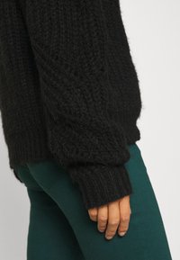 Czarny sweter z dzianiny o teksturowanym wykończeniu, z prążkowanymi mankietami. W połączeniu z zielonymi spodniami, pokazujący zbliżenie na rękaw i dłoń.