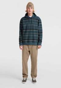 Capuchon sweatshirt in donker teal met horizontale strepen, gecombineerd met beige broek en grijze schoenen. De beanie heeft een logo patch.