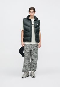 Fiatal férfi áll fekete pufi mellényben, világos mintás pólóban, fekete-fehér mintás bő nadrágban, szürke sportcipőben, jobb kezében fekete sapkát tartva.