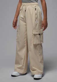 Beige cargo pants with elastic waistband, two side pockets, and one back pocket. Made from lightweight, textured material. Logo accent near waist.