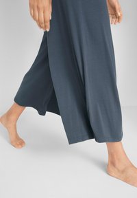 Abito lungo di un grigio scuro con una vestibilità rilassata. Tessuto morbido, ampi spacchi laterali e un orlo liscio e rifinito. Indossato a piedi nudi, evidenziando uno stile casual.