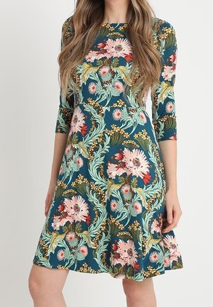 Femme portant une robe bleu sarcelle longueur genou avec des manches trois-quarts et un grand motif floral rose et vert.