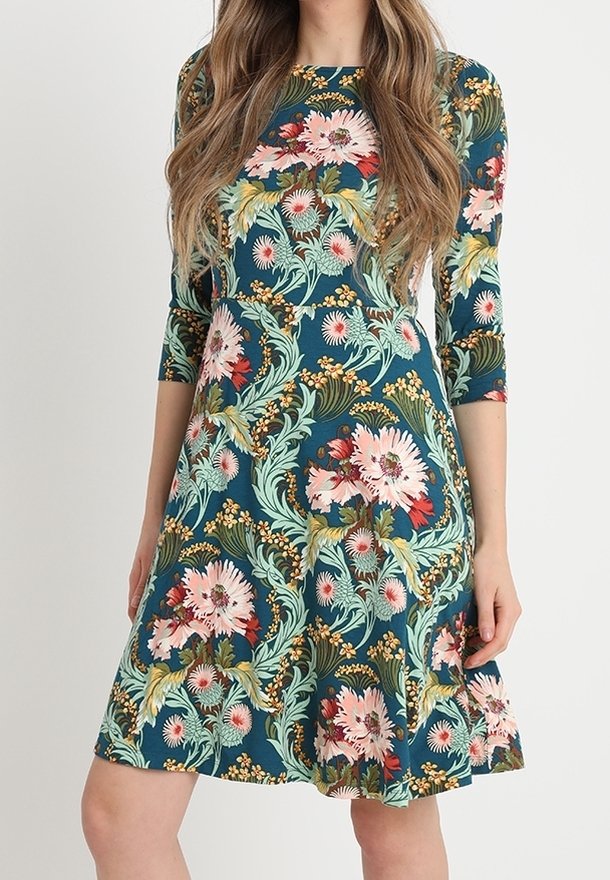 Femme portant une robe bleu sarcelle longueur genou avec des manches trois-quarts et un grand motif floral rose et vert.