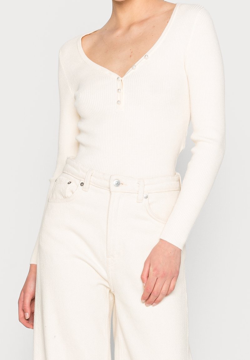 Vitt ribbat långärmad topp med V-ringning och knapprad vid halslinjen, i kombination med högmidjade vita vida byxor.