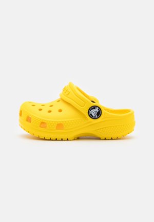 Lys gul slip-on træsko med ventilationshuller, en struktureret sål og en justerbar rem med en cirkulær logo på siden.