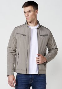 Chaqueta bomber gris claro con cuello alzado, dos bolsillos en el pecho con cremallera y acentos acolchados en los hombros. Combinada con una camiseta blanca.
