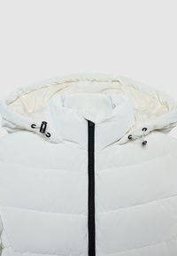 Doudoune blanche avec un col montant et une capuche à cordon de serrage ; équipée d'une fermeture éclair noire à l'avant et d'une doublure intérieure douce.