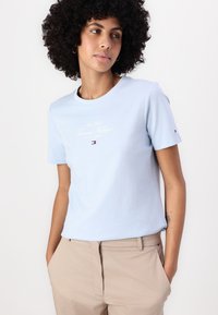 T-shirt bleu clair à manches courtes en coton doux, avec un logo en script doré et un accent logo sur la manche.